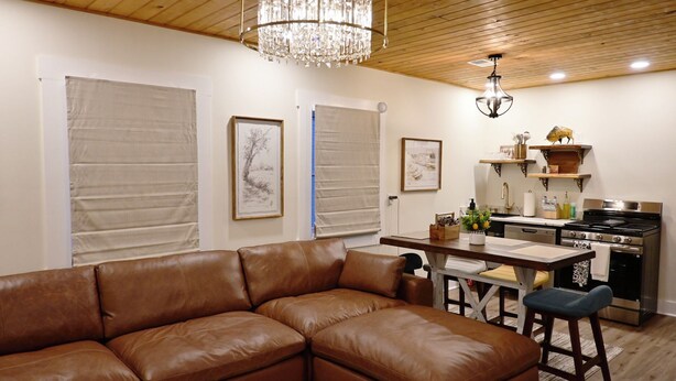 A cozy living room with a brown leather sofa, a wooden dining table, and a kitchen area with a stove and shelves. - Alterra:Slees 8,Steps to Main St,Serene (Madison)