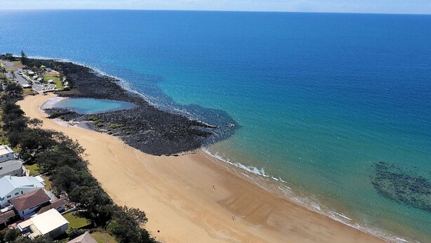 An aerial view of a beach with clear blue waters, a sandy shore, and a rocky outcrop.