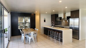A modern kitchen with a central island, white chairs, dark cabinetry, and a stainless steel refrigerator.