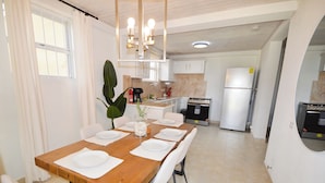 Un coin repas avec une table en bois dressée pour quatre personnes, des chaises blanches, un vase avec des plantes vertes, et, au fond, une cuisine équipée d’un réfrigérateur et d’une cuisinière.