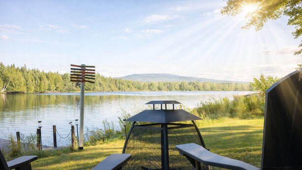 Et udendørs siddeområde ved søen med en pavillon, picnicborde og et vejskilt.
