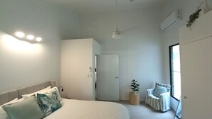 Ein modernes Schlafzimmer mit Bett, Nachttisch und Stuhl. Eine Tür führt in einen anderen Raum, eine Zimmerpflanze und ein Fenster mit Jalousien.