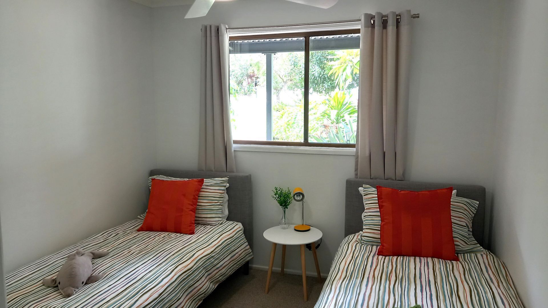 A small, clean room with two single beds, each with striped bedding and red pillows, a small round table with a vase, and a window with curtains.