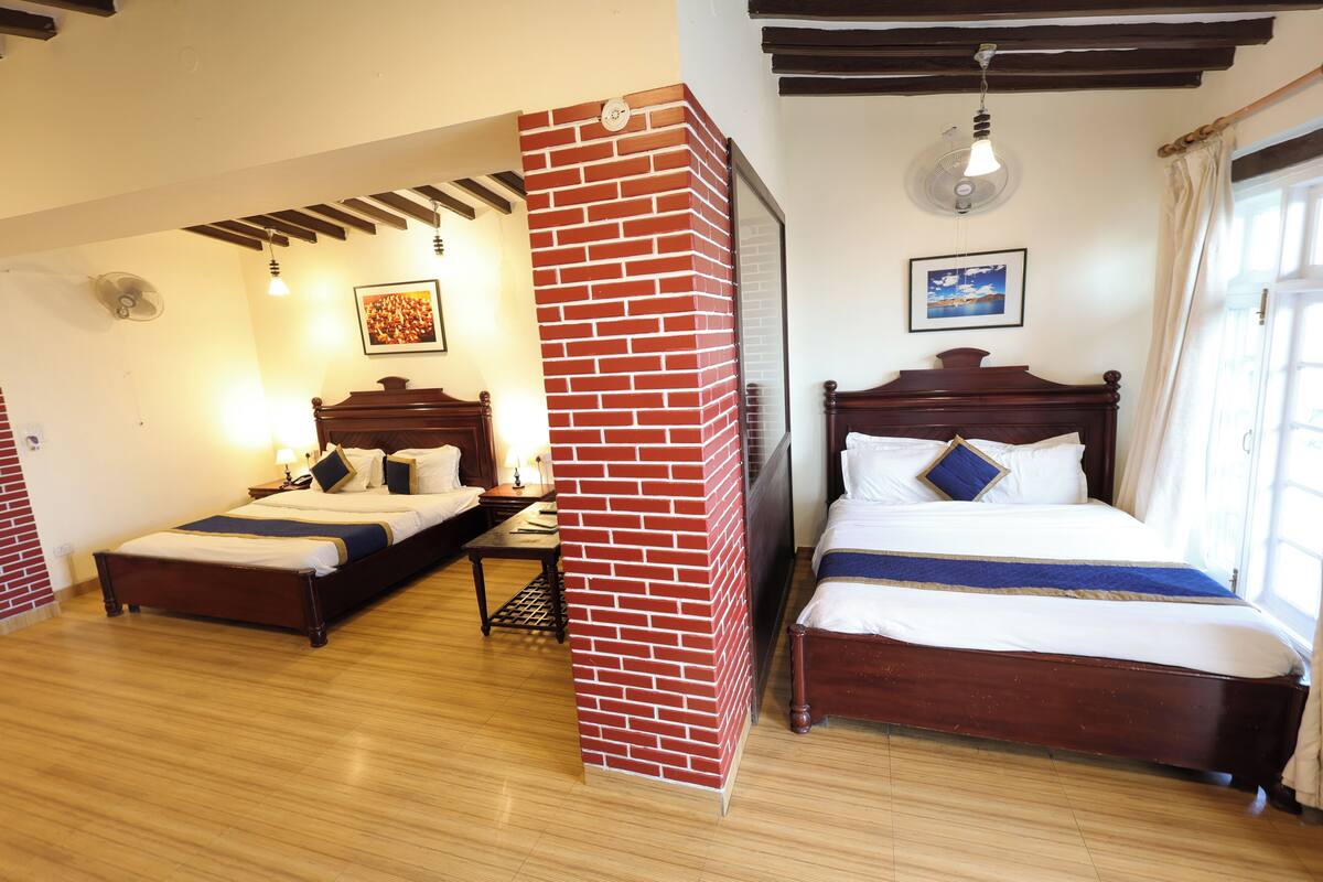 Een hotelkamer met twee houten bedden, een accentmuur van baksteen, een plafondventilator en een raam met gordijnen.