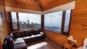 A wooden cabin room with a leather sofa, a small bedside table with a lamp, and large windows offering a mountain view. - Sansa By The Woods - Manor Mussoorie (Mussoorie)