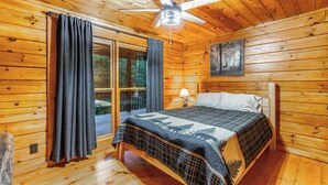 A wooden cabin bedroom with a bed, a ceiling fan, a window with curtains, and a painting on the wall.
