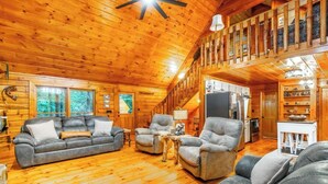 A log cabin interior with a wooden ceiling, a sofa, armchairs, a dining table, and a kitchen area.