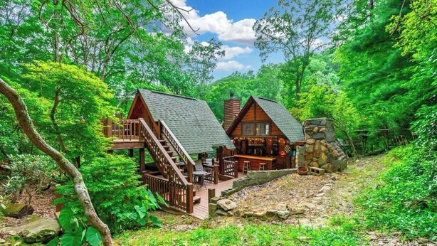 A cabin with a wooden deck, outdoor seating, and a stone fireplace.