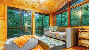 A cozy cabin interior with a wooden floor, a built-in bench with cushions, and large windows offering a view of the forest.