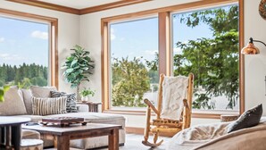 A cozy living room with a sofa, a rocking chair, and a coffee table. Large windows provide a view of greenery outside.