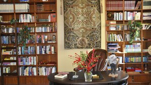 Een bibliotheek met houten boekenkasten vol boeken, een decoratief wandtapijt en een bureau met een vaas rode bessen.