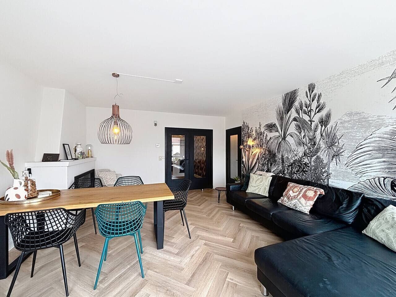 A modern living room with a wooden dining table, black and turquoise chairs, a black sofa with patterned cushions, and a large wall mural.