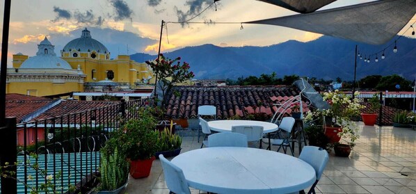 Una terraza en la azotea con mesas al aire libre, plantas en macetas y vistas a un edificio amarillo con cúpula y montañas al fondo.