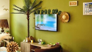 A room with a green wall, a mounted TV displaying a beach scene, a palm tree, and a small table with decorative items.