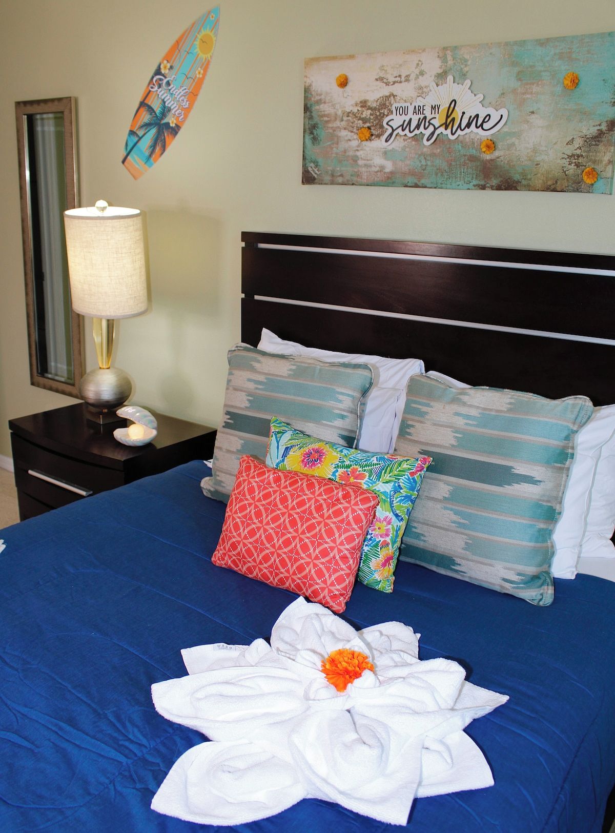 Un lit bien fait, avec une couvre-lit bleue, plusieurs oreillers, une serviette décorative, une table de chevet avec une lampe et un élément décoratif mural.