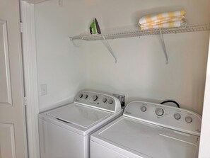 A laundry room with two front-loading washers and dryers, a metal shelf with a towel, and a power outlet on the wall.