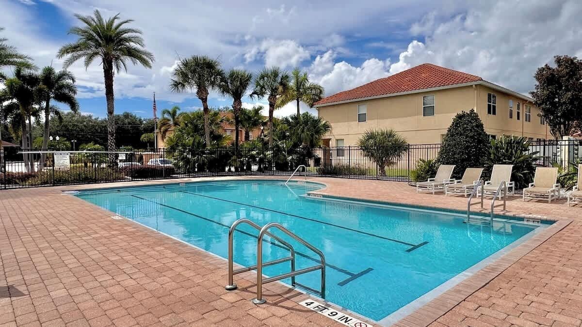 A swimming pool with a metal ladder, surrounded by a brick patio, palm trees, and a building with a red roof.