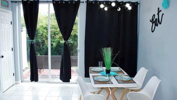 Un coin repas avec une table dressée pour quatre personnes, des chaises blanches, des rideaux noirs et un panneau mural.