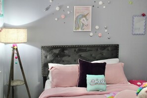 A bedroom with a tufted headboard, a bed with pink bedding, a teal pillow with the word 'Chill,' and a wall decoration of a unicorn.