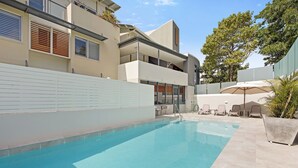 Un espace piscine moderne avec une piscine à l’eau d’un bleu limpide, des chaises longues et un mur blanc orné de lattes horizontales.