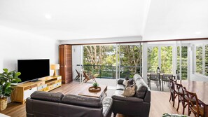 Ein modernes Wohnzimmer mit Sofa, Fernseher, Couchtisch aus Holz und einem Balkon mit Blick auf Bäume.