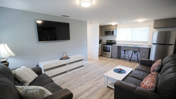 Una sala de estar moderna con un televisor de pantalla plana, una consola blanca y un sofá. La zona de la cocina incluye un microondas, horno y refrigerador.