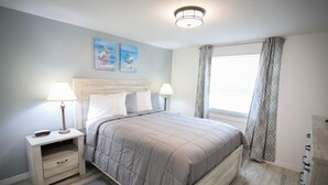 A bedroom with a bed, two bedside tables, a nightstand, a lamp, a window with curtains, and two wall art pieces. - Seashell Room Manasota Delux Villas (Sarasota County)