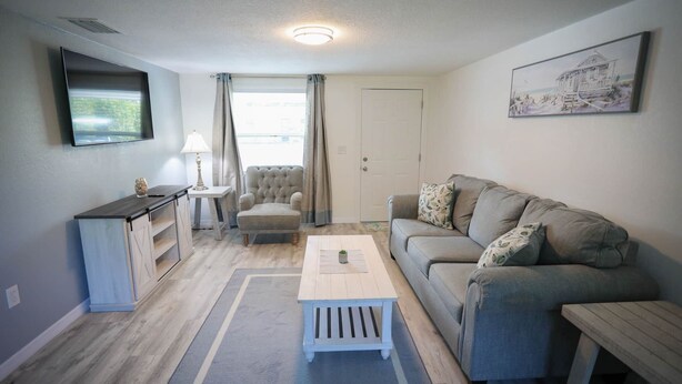 A modern living room with a grey sofa, a white coffee table, a grey armchair, a small side table with a lamp, and a TV mounted on the wall. - Seashell Room Manasota Delux Villas (Sarasota County)