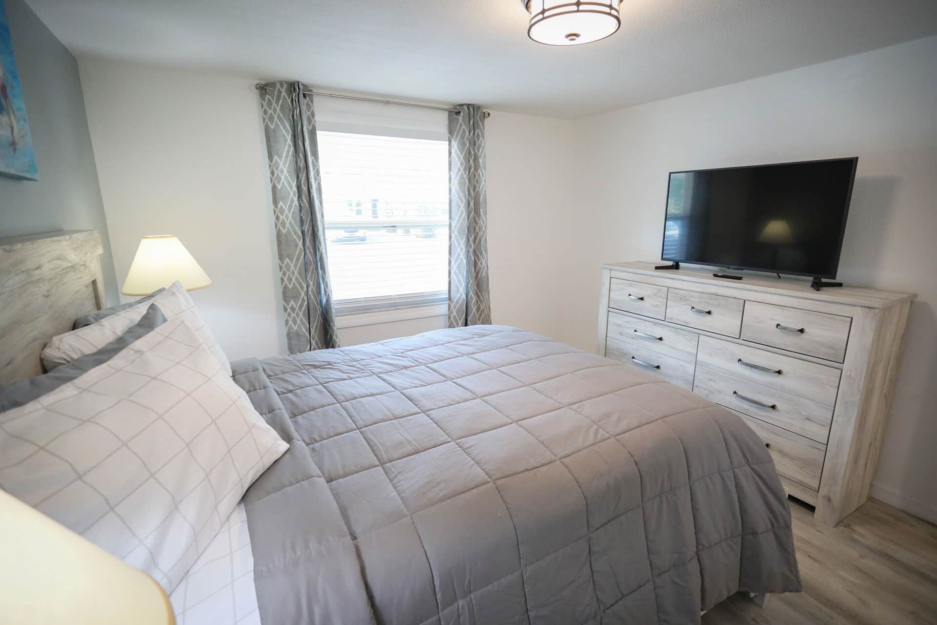 Een slaapkamer met een bed, een televisie op een dressoir en een raam met gordijnen.