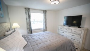 A bedroom with a bed, a TV on a dresser, and a window with curtains. - Seashell Room Manasota Delux Villas (Sarasota County)