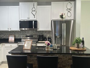 A modern kitchen with white cabinets, stainless steel appliances, a granite countertop, and a small island with chairs. - Spacious 4-bedroom house in the heart of Oakleaf (Orange Park)