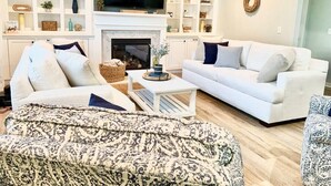 A living room with a fireplace, white sofas, a patterned ottoman, and a TV mounted above the fireplace. - Saltee Cove Coastal Getaway - Charming 3 Bedroom/2.5 Bath Lowcountry Cottage (Bluffton)