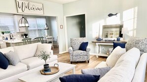 A modern living room with a sofa, armchairs, and a coffee table. A kitchen is visible in the background with a dining area. - Saltee Cove Coastal Getaway - Charming 3 Bedroom/2.5 Bath Lowcountry Cottage (Bluffton)