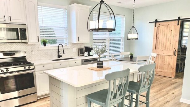 A modern kitchen with white cabinets, stainless steel appliances, a central island with bar stools, and a wooden barn-style door. - Saltee Cove Coastal Getaway - Charming 3 Bedroom/2.5 Bath Lowcountry Cottage (Bluffton)