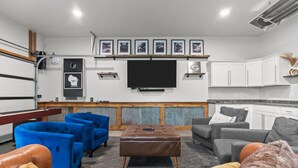 Un salon doté d’un téléviseur à écran plat, de canapés bleus et gris, d’un pouf marron et d’un coin cuisine avec des meubles blancs.