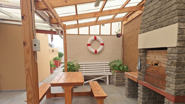 An outdoor patio area with wooden benches, a table, potted plants, a lifebuoy, and a stone fireplace. - Spacious cosy, self- Catering loft with own entrance in safe & secure area. (Swakopmund)