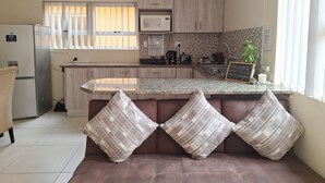 A modern kitchen with a built-in island, a refrigerator, a microwave, and a sink. There are striped pillows on a bench in front of the kitchen. - Spacious cosy, self- Catering loft with own entrance in safe & secure area. (Swakopmund)