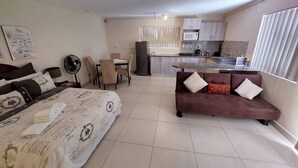 A compact living space with a bed, a sofa, a dining table with chairs, a kitchenette, and a fan. - Spacious cosy, self- Catering loft with own entrance in safe & secure area. (Swakopmund)