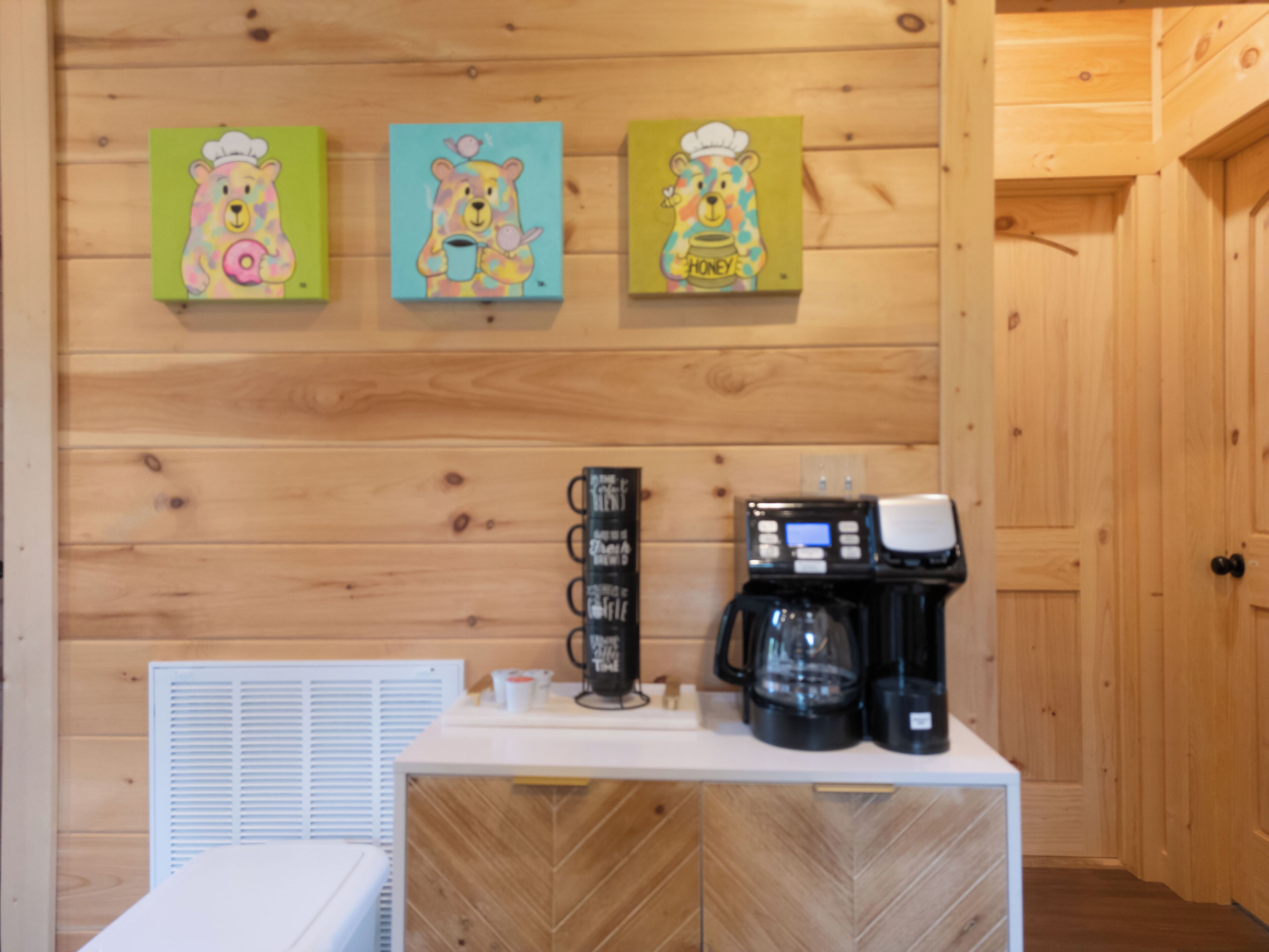 A kitchen area with wooden walls, a white countertop, a coffee maker, and three bear-themed paintings.