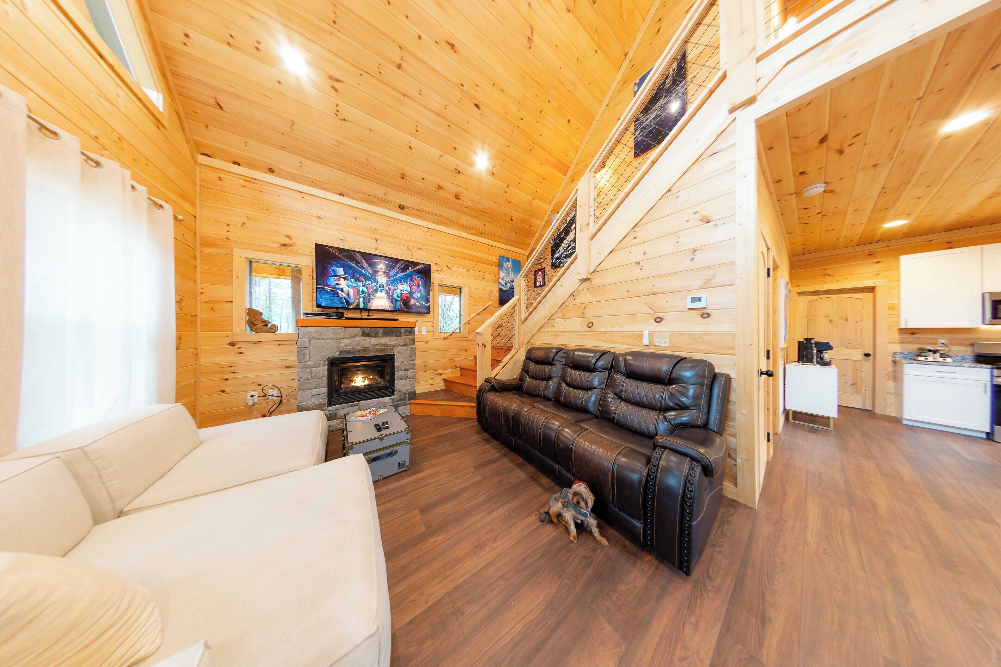 A wooden cabin interior with a living room, a fireplace, a leather sofa, and a staircase leading to another level.