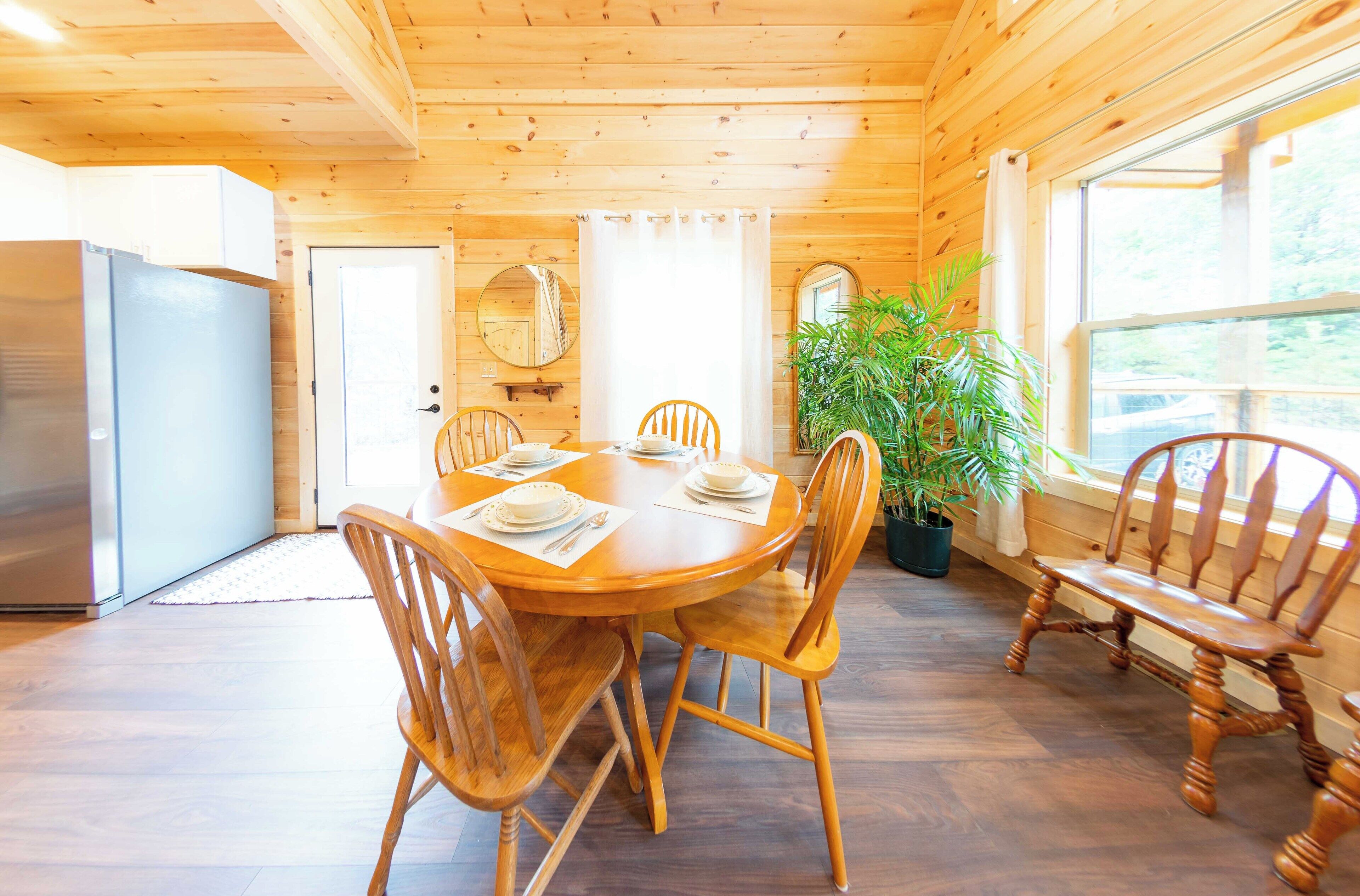 Un interno in legno con un tavolo da pranzo apparecchiato per quattro persone, circondato da sedie in legno. Nell'angolo si trova una pianta in vaso e un grande specchio è appeso alla parete.