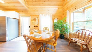 Interior de madera con una mesa de comedor preparada para cuatro personas, rodeada de sillas de madera. Una planta en maceta está en la esquina, y un espejo grande cuelga en la pared.