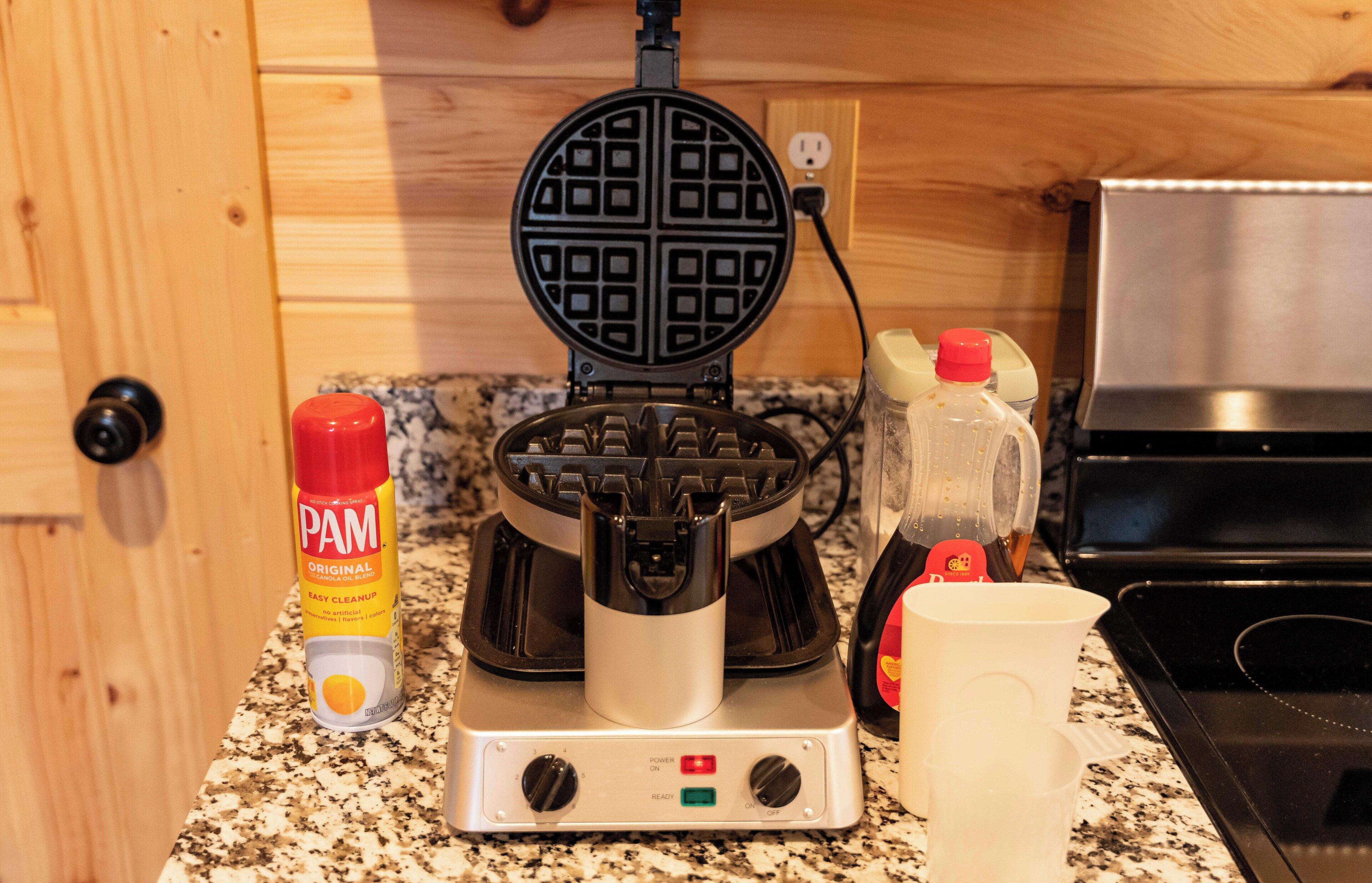 A waffle maker on a kitchen counter with a bottle of PAM oil, a container of oil, and a cup.