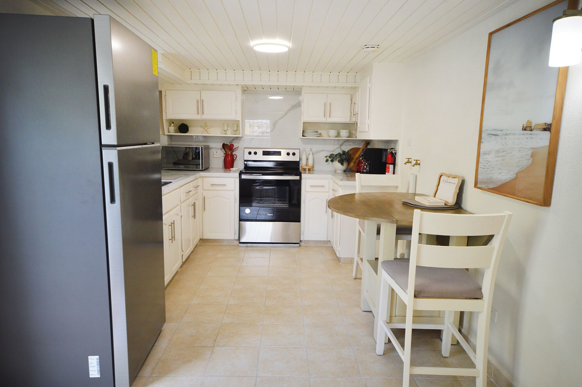 A compact kitchen with white cabinets, a stainless steel refrigerator, a built-in oven, and a dining area with a wooden table and white chairs.