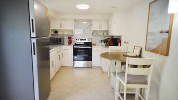 A compact kitchen with white cabinets, a stainless steel refrigerator, a built-in oven, and a dining area with a wooden table and white chairs.