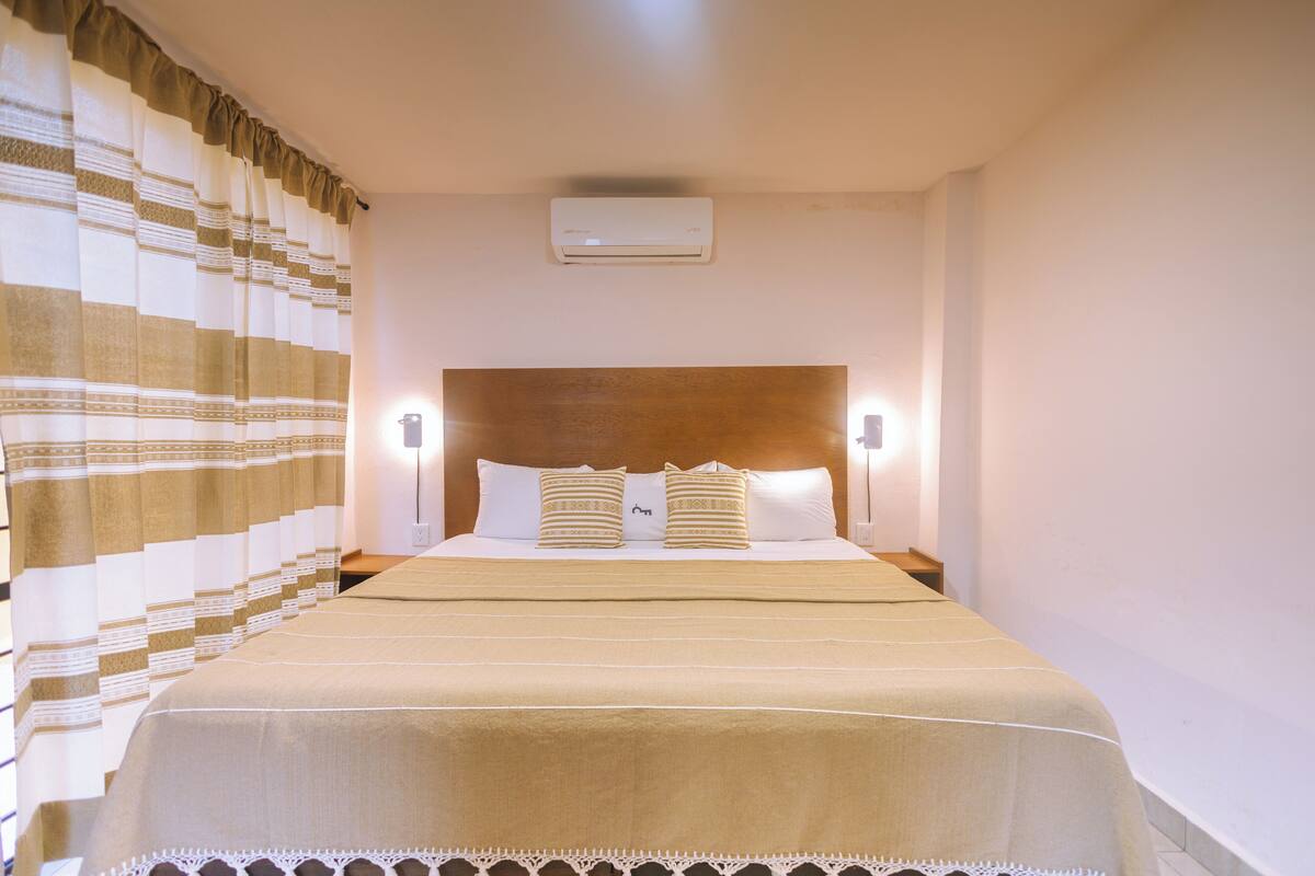 Una cama bien hecha con cabecera de madera, dos lámparas de noche y una cortina estampada.