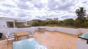 Un patio al aire libre con una mesa y sillas de vidrio, una mesita auxiliar y vistas a un edificio histórico al fondo.