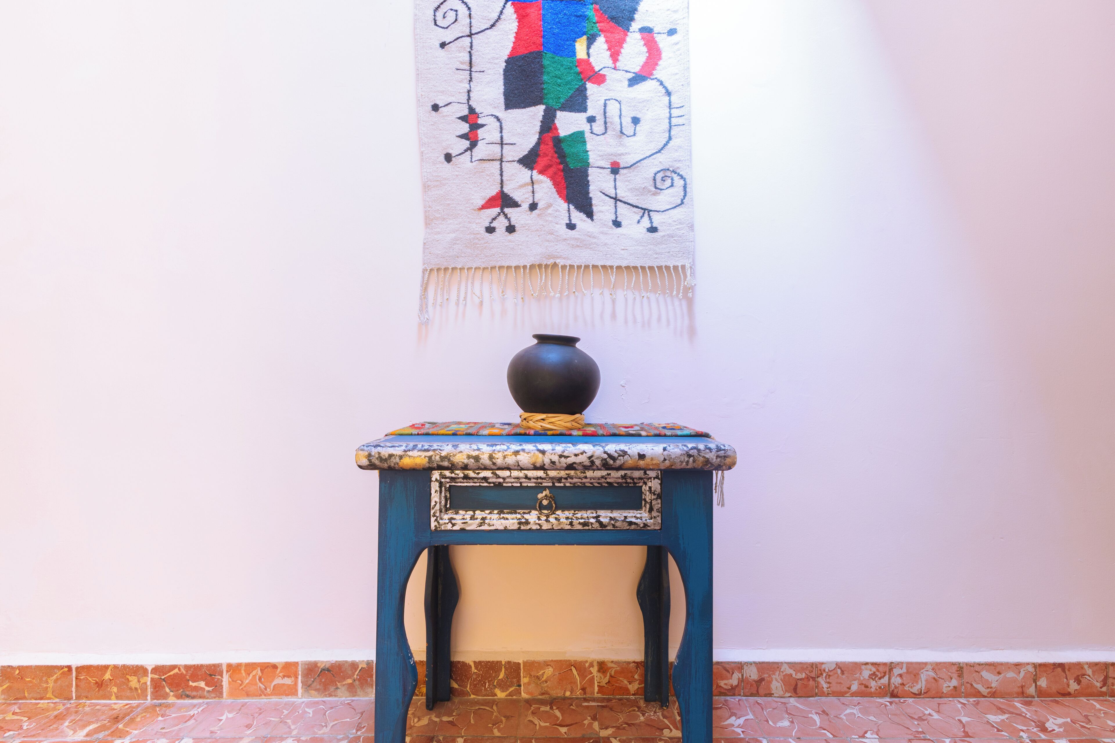 Una mesa azul con una superficie decorativa y un florero negro encima, frente a una pared con un tapiz colorido.