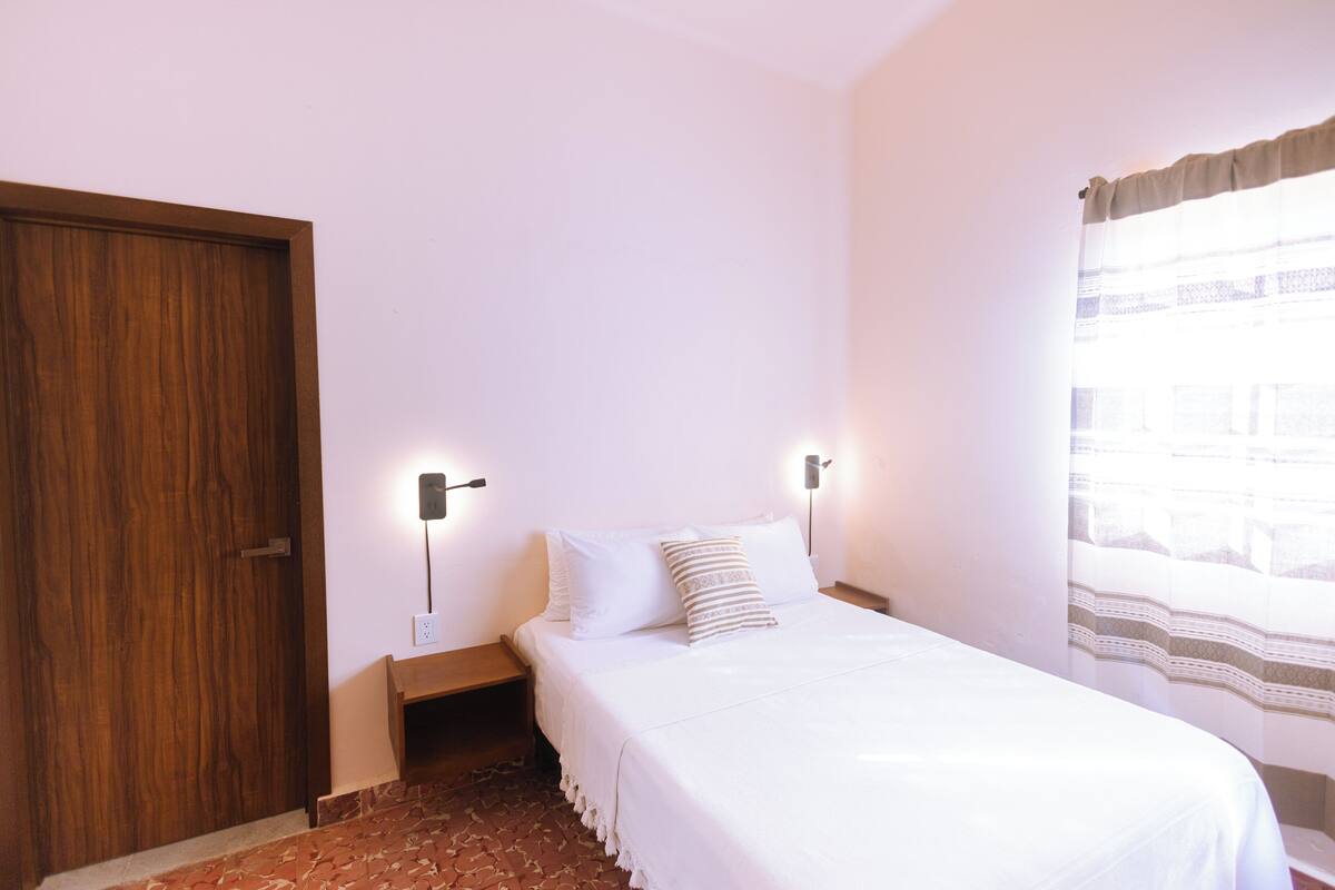 Un dormitorio con puerta de madera, cama con ropa blanca, dos lámparas de noche y una ventana con cortinas.