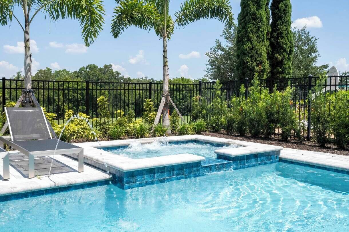 A swimming pool with a hot tub, surrounded by a fence and lush greenery.
